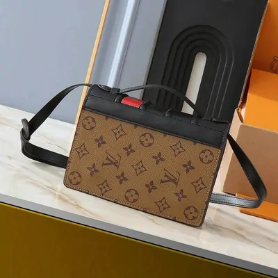 A Louis Vuitton crossbody bag in the monogram pattern, standing on a pale stone surface with its orange box behind it.